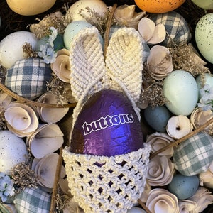 May include: A crocheted bunny basket containing a purple chocolate egg with the word "buttons" on it. The basket is surrounded by decorative eggs and other Easter decorations.