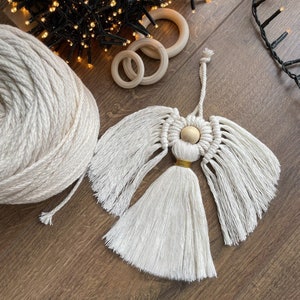 May include: A white macrame angel ornament with a wooden bead and gold accents. The angel has a long tassel and is hanging from a string.