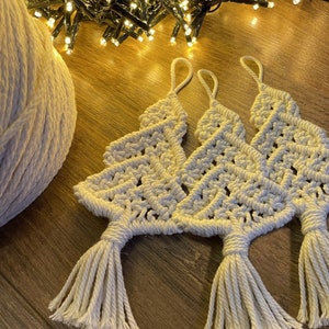 May include: Cream-coloured macrame Christmas tree decorations with a looped top and tassel base. The ornaments are displayed on a wooden surface with a ball of cream-coloured yarn and a string of fairy lights.
