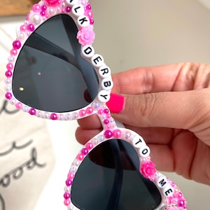 May include: White heart-shaped sunglasses with pink and white beads and the words "TALK TO ME" and "DERBY" spelled out on the frames.