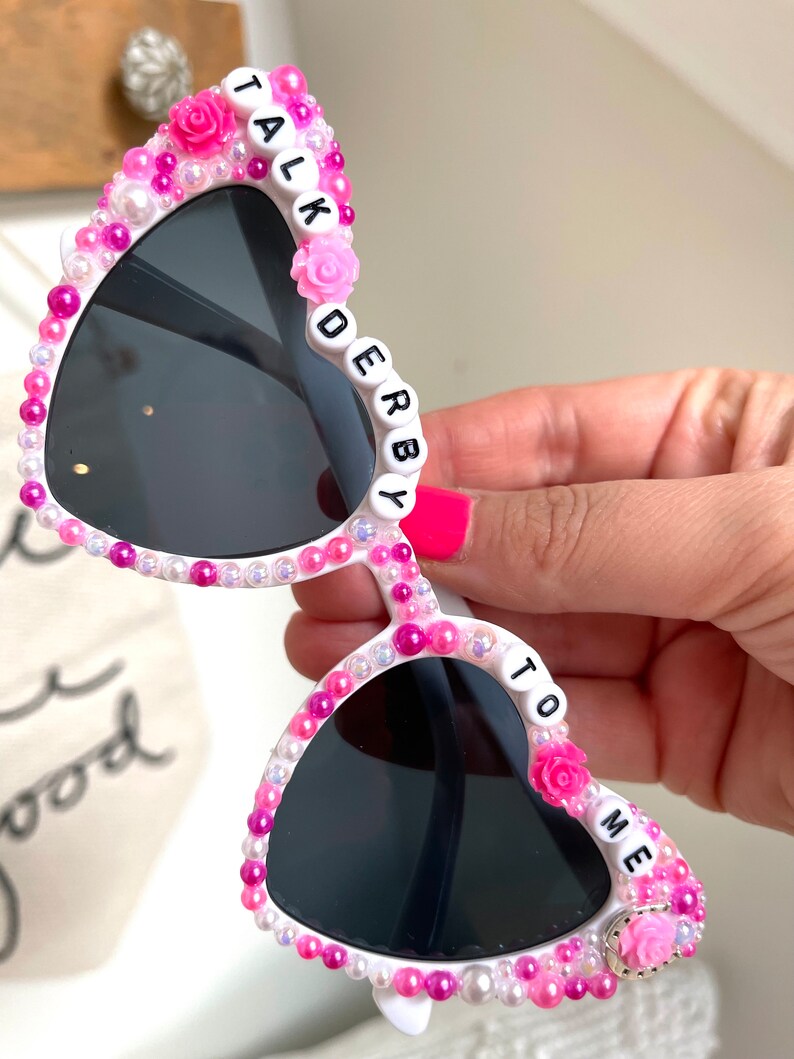 May include: White heart-shaped sunglasses with pink and white beads and the words "TALK TO ME" and "DERBY" spelled out on the frames.