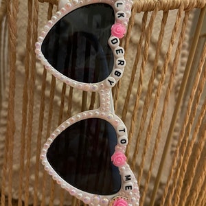May include: White heart-shaped sunglasses with black lenses and pink flowers. The sunglasses have white beads and the words "TALK DERBY TO ME" spelled out on the frames.
