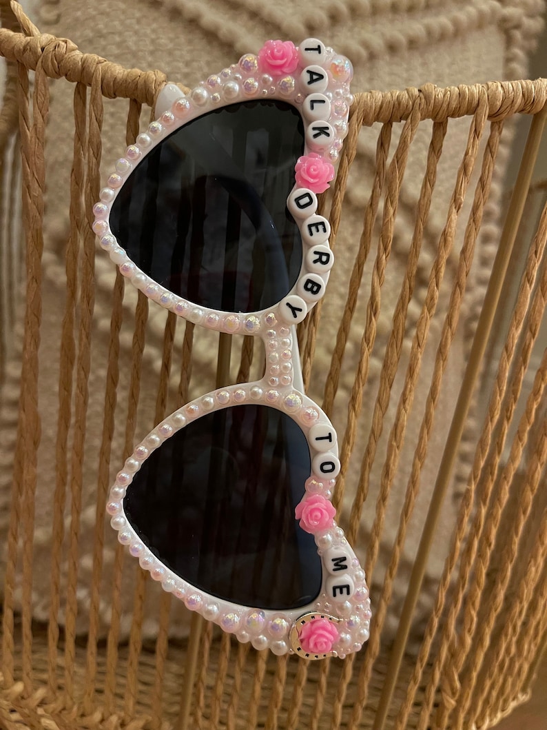 May include: White heart-shaped sunglasses with black lenses and pink flowers. The sunglasses have white beads and the words "TALK DERBY TO ME" spelled out on the frames.
