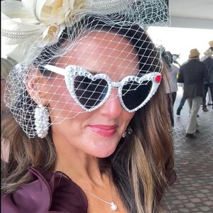 May include: A woman wearing a white hat with a veil and pearl embellishments, heart-shaped sunglasses with rhinestones and the words "RIDIN DERBY" on the frames, a pearl necklace, and a dark purple one-shoulder dress.