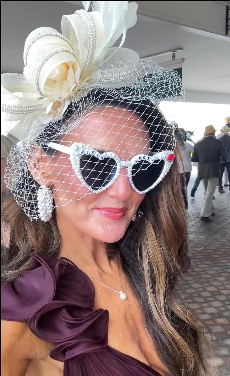 May include: A woman wearing a white hat with a veil and pearl embellishments, heart-shaped sunglasses with rhinestones and the words "RIDIN DERBY" on the frames, a pearl necklace, and a dark purple one-shoulder dress.