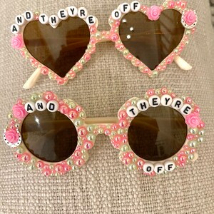 May include: Two pairs of heart-shaped and round sunglasses with pink and white beads and the words "And They're Off" on the frames.
