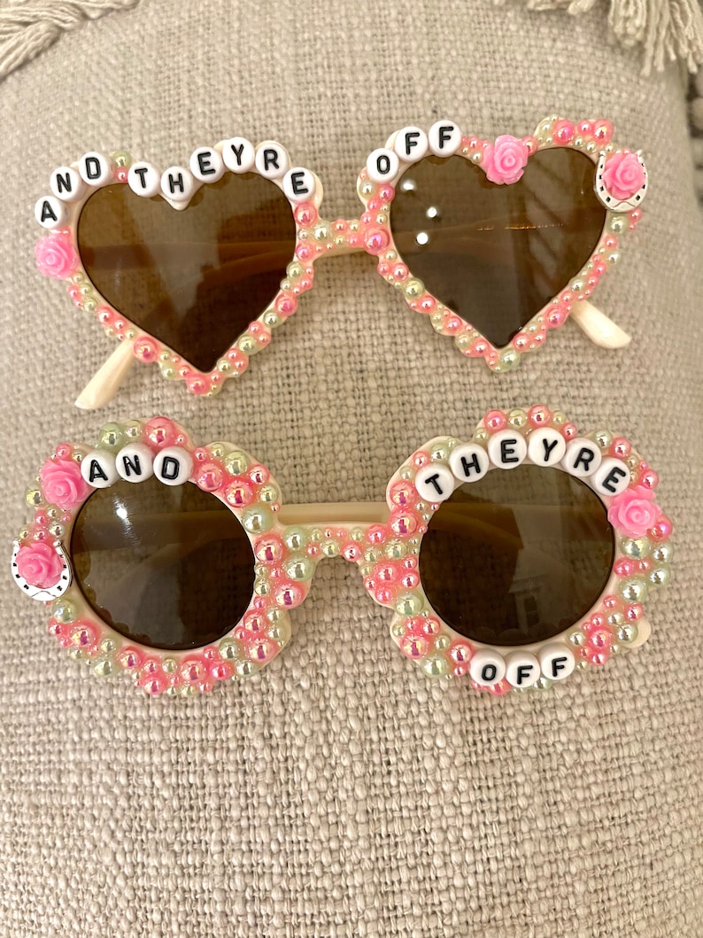 May include: Two pairs of heart-shaped and round sunglasses with pink and white beads and the words "And They're Off" on the frames.