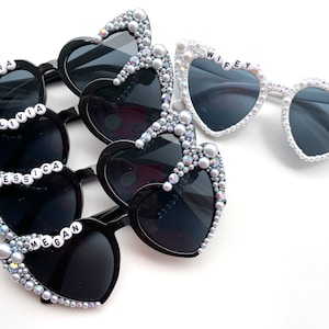 May include: Set of heart-shaped sunglasses with pearl and rhinestone embellishments. Four black sunglasses have the names "Sara", "Olivia", "Jessica", and "Megan" on the frames. One white sunglass has the word "WIFEY" on the frame.