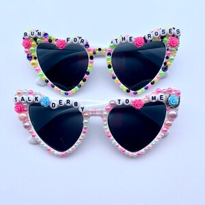 May include: White heart-shaped sunglasses with black lenses and pearl and flower embellishments. The sunglasses have the words "Run for the Roses" and "Talk Derby to Me" on them.