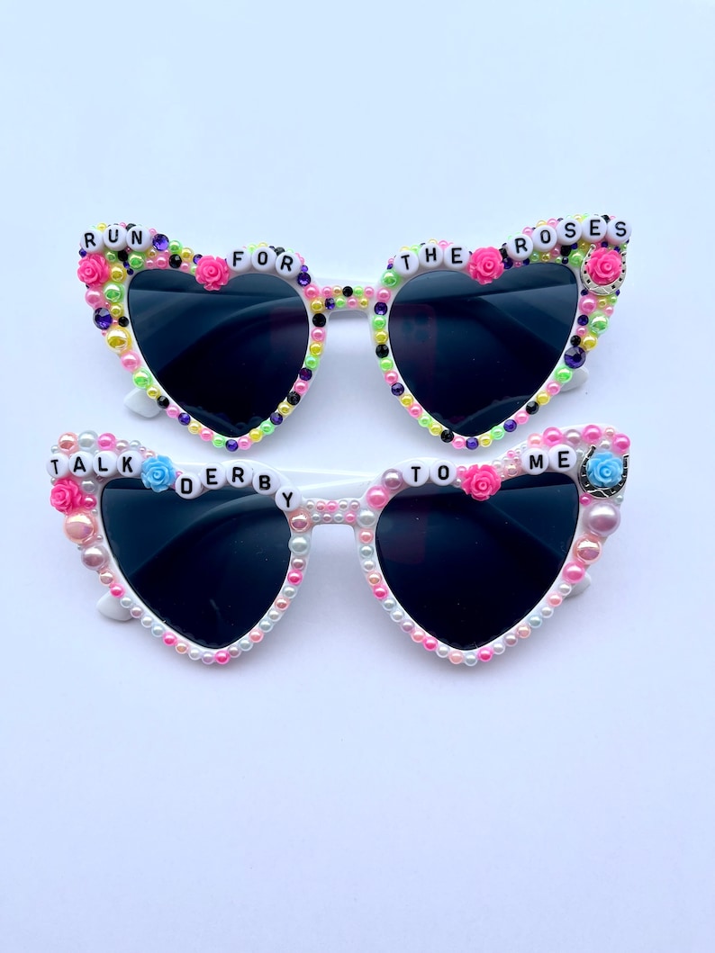 May include: White heart-shaped sunglasses with black lenses and pearl and flower embellishments. The sunglasses have the words "Run for the Roses" and "Talk Derby to Me" on them.