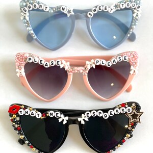 Op de afbeelding: Drie paar hartvormige zonnebrillen met strass en decoratieve versieringen. Het bovenste paar is lichtblauw met de woorden "SHORT N SWEET TOUR" op de monturen. Het middelste paar is roze met de woorden "PLEASE PLEASE" op de monturen. Het onderste paar is zwart met de woorden "THAT ME ESPRESSO" op de monturen.