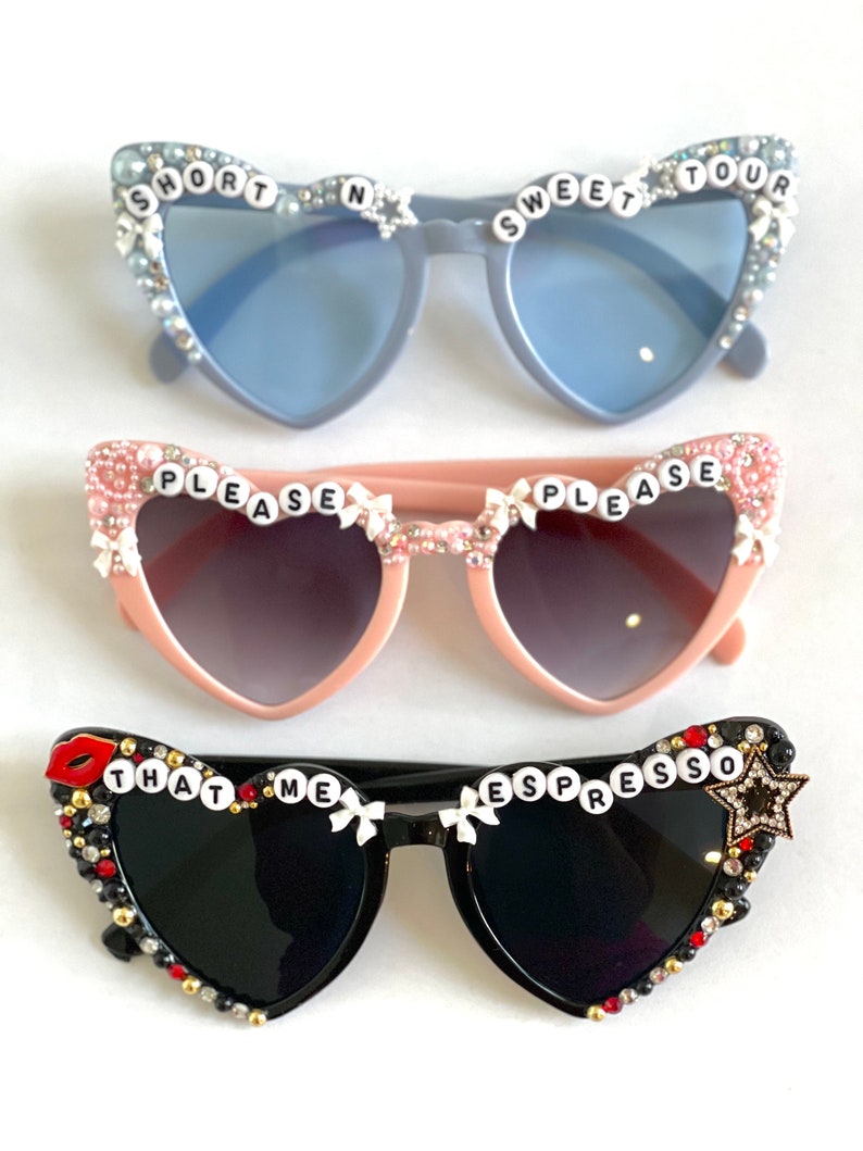 Op de afbeelding: Drie paar hartvormige zonnebrillen met strass en decoratieve versieringen. Het bovenste paar is lichtblauw met de woorden "SHORT N SWEET TOUR" op de monturen. Het middelste paar is roze met de woorden "PLEASE PLEASE" op de monturen. Het onderste paar is zwart met de woorden "THAT ME ESPRESSO" op de monturen.