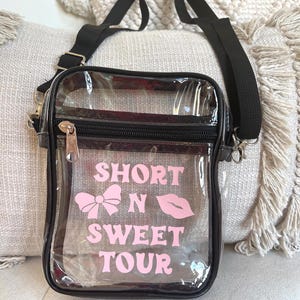 Op de afbeelding: Transparante crossbody tas met een verstelbare zwarte schouderband. De tas heeft een ritssluiting en heeft roze tekst die "Short N Sweet Tour" zegt met een roze strik en lippen graphic.