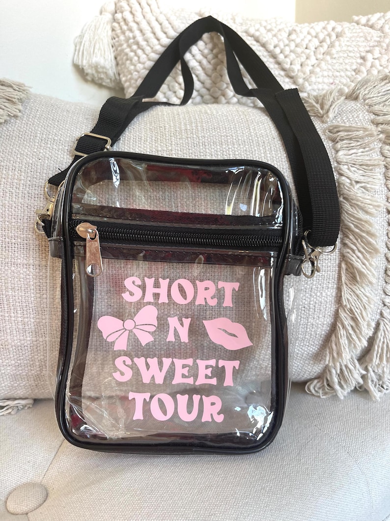 Op de afbeelding: Transparante crossbody tas met een verstelbare zwarte schouderband. De tas heeft een ritssluiting en heeft roze tekst die "Short N Sweet Tour" zegt met een roze strik en lippen graphic.