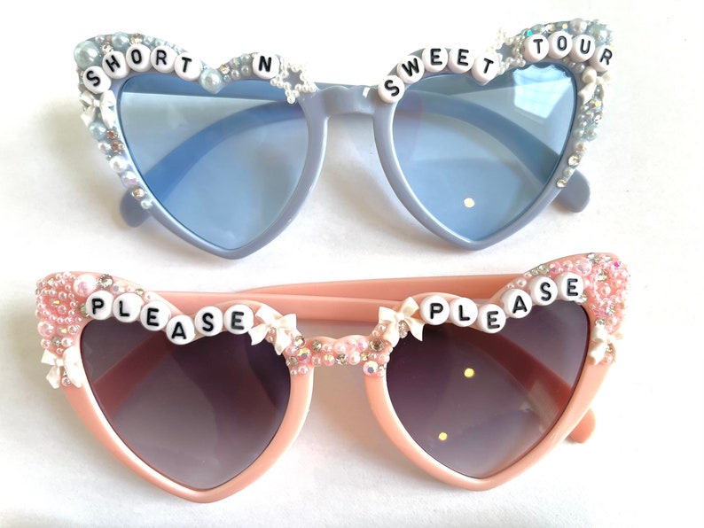 Op de afbeelding: Twee paar hartvormige zonnebrillen met strass en strikjes. Het bovenste paar is lichtblauw met de woorden "SHORT N SWEET TOUR" op de monturen. Het onderste paar is roze met de woorden "PLEASE PLEASE" op de monturen.