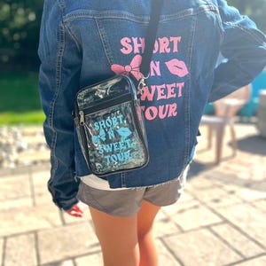 Op de afbeelding: Een persoon draagt een blauw denim jack met de tekst "Short N Sweet Tour" bedrukt op de rug. Ze dragen ook een doorzichtige crossbody tas van plastic met dezelfde tekst bedrukt op de voorkant.