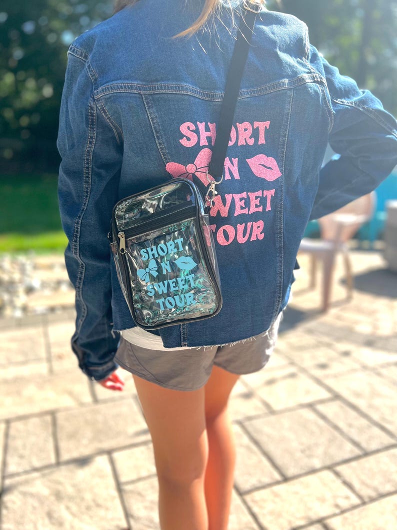 Op de afbeelding: Een persoon draagt een blauw denim jack met de tekst "Short N Sweet Tour" bedrukt op de rug. Ze dragen ook een doorzichtige crossbody tas van plastic met dezelfde tekst bedrukt op de voorkant.