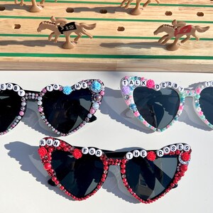 May include: Three pairs of heart-shaped sunglasses with black lenses and beaded embellishments. The sunglasses are decorated with colorful beads and letters spelling out "Talk Derby To Me" and "Run For The Roses".