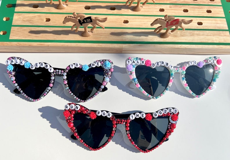 May include: Three pairs of heart-shaped sunglasses with black lenses and beaded embellishments. The sunglasses are decorated with colorful beads and letters spelling out "Talk Derby To Me" and "Run For The Roses".