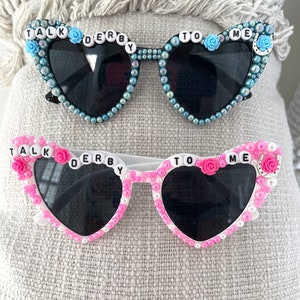 May include: Two pairs of heart-shaped sunglasses with rhinestones and the words "Talk Derby To Me" on the frames. One pair is black with blue rhinestones and the other is white with pink rhinestones.