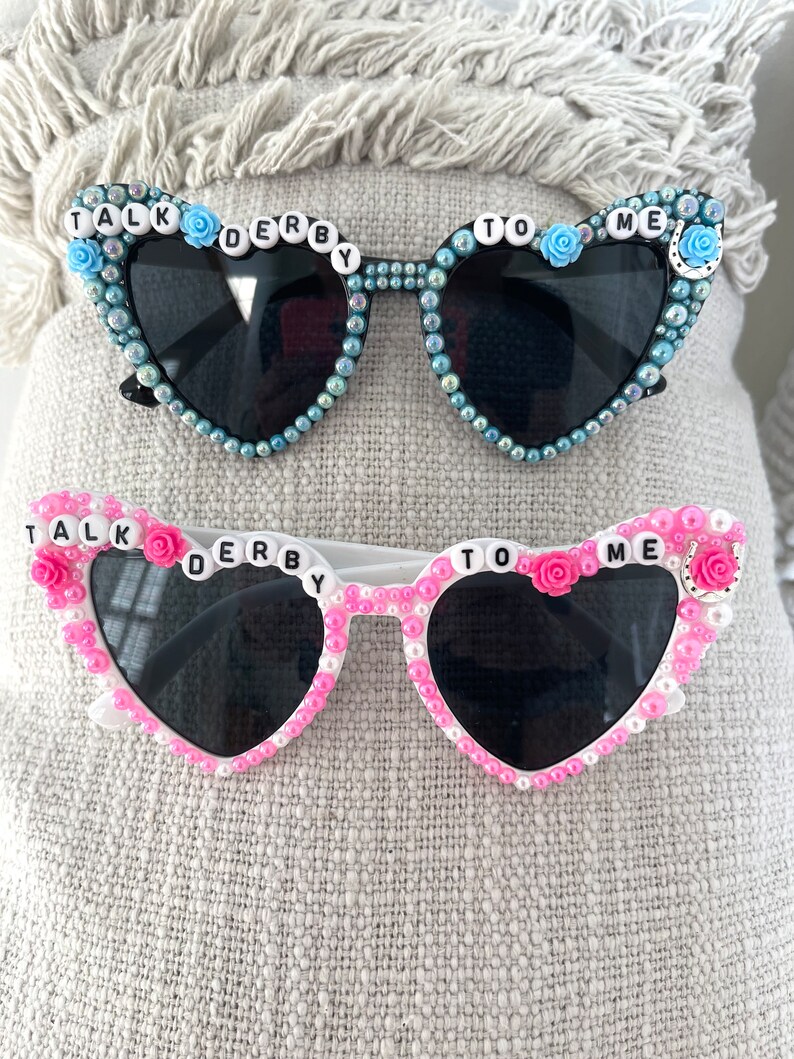 May include: Two pairs of heart-shaped sunglasses with rhinestones and the words "Talk Derby To Me" on the frames. One pair is black with blue rhinestones and the other is white with pink rhinestones.