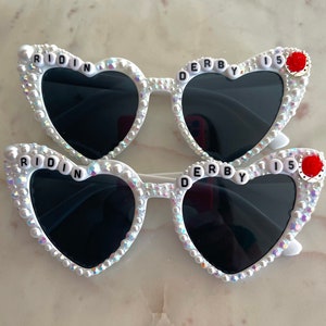 May include: White heart-shaped sunglasses with black lenses and rhinestones. The sunglasses have the words "RIDIN' DERBY 15" on the frames.