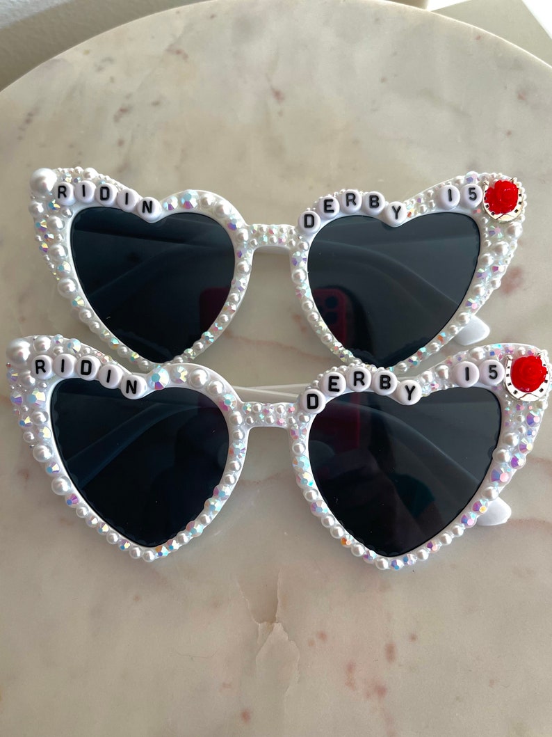 May include: White heart-shaped sunglasses with black lenses and rhinestones. The sunglasses have the words "RIDIN' DERBY 15" on the frames.