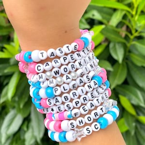 Op de afbeelding: Een stapel kleurrijke kralenarmbanden met woorden zoals "Taste", "Carpenter", "Working", "Mass", "Please", "Sabrina", "Espresso", "Feather", "Short" en "Sc" erop. Elke armband heeft een stervormige bedel.