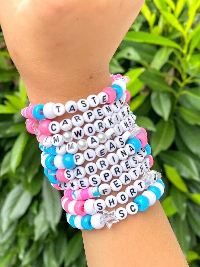 Op de afbeelding: Een stapel kleurrijke kralenarmbanden met woorden zoals "Taste", "Carpenter", "Working", "Mass", "Please", "Sabrina", "Espresso", "Feather", "Short" en "Sc" erop. Elke armband heeft een stervormige bedel.