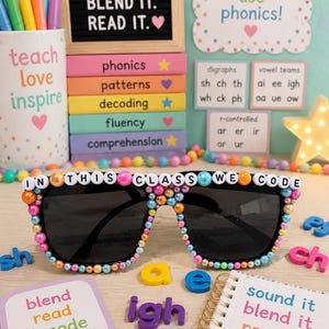 May include: Black sunglasses decorated with colorful beads and white letter beads spelling out IN THIS CLASS WE CODE. The frame is adorned with a mix of pink, blue, orange, and yellow beads, creating a vibrant, textured border around the dark lenses.