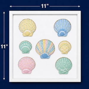 May include: A square white frame containing seven decorative seashells arranged on a white background. The shells are painted in pastel yellow, blue, pink, and green with white polka dot patterns. The frame measures 11 inches by 11 inches.