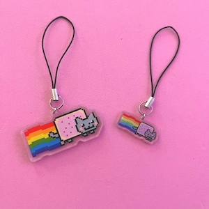 May include: Two Nyan Cat charms with black cords and silver clasps. The charms depict pixelated images of a grey cat with a rainbow trail, one large and one small, against a pink backdrop.