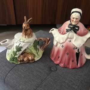 May include: Two ceramic teapots, one shaped like a rabbit in a burrow and the other shaped like a woman in a pink dress. Both teapots have hand-painted details.