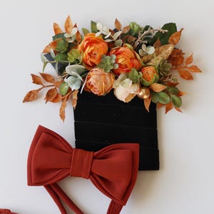 May include: A rust-colored bow tie with a long strap. The bow tie is made of a soft fabric and has a classic shape. The bow tie is lying on a white surface.