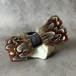 Puede incluir: Una pajarita decorativa hecha de plumas marrones y blancas con una banda de cuero negro. Las plumas tienen un patrón y una textura naturales. La pajarita se exhibe sobre una piedra blanca sobre un fondo gris.