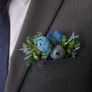 May include: A charcoal gray suit jacket with a white collared shirt and a dark blue tie. A floral pocket square accessory featuring blue peonies, small blue flowers, and green foliage is tucked into the breast pocket of the suit jacket.