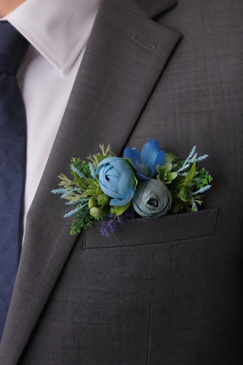 May include: A charcoal gray suit jacket with a white collared shirt and a dark blue tie. A floral pocket square accessory featuring blue peonies, small blue flowers, and green foliage is tucked into the breast pocket of the suit jacket.
