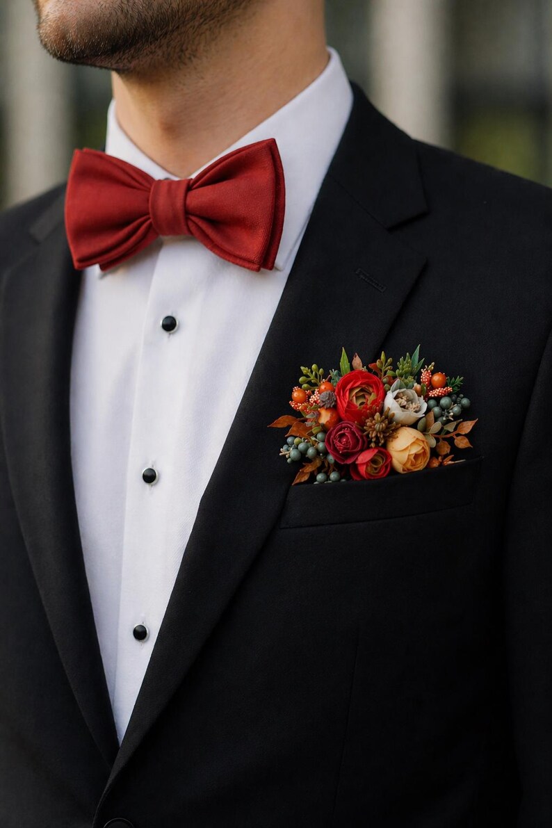 May include: A black suit jacket worn over a white dress shirt with black buttons. The outfit features a solid dark red bow tie and a pocket square arrangement consisting of artificial red, orange, and yellow flowers with green foliage.