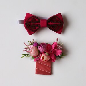 May include: A dark pink fabric bow tie featuring a dark brown laser-cut wooden overlay with intricate scrollwork, paired with a matching boutonniere composed of artificial pink and peach flowers, green foliage, and a rectangular fabric base.