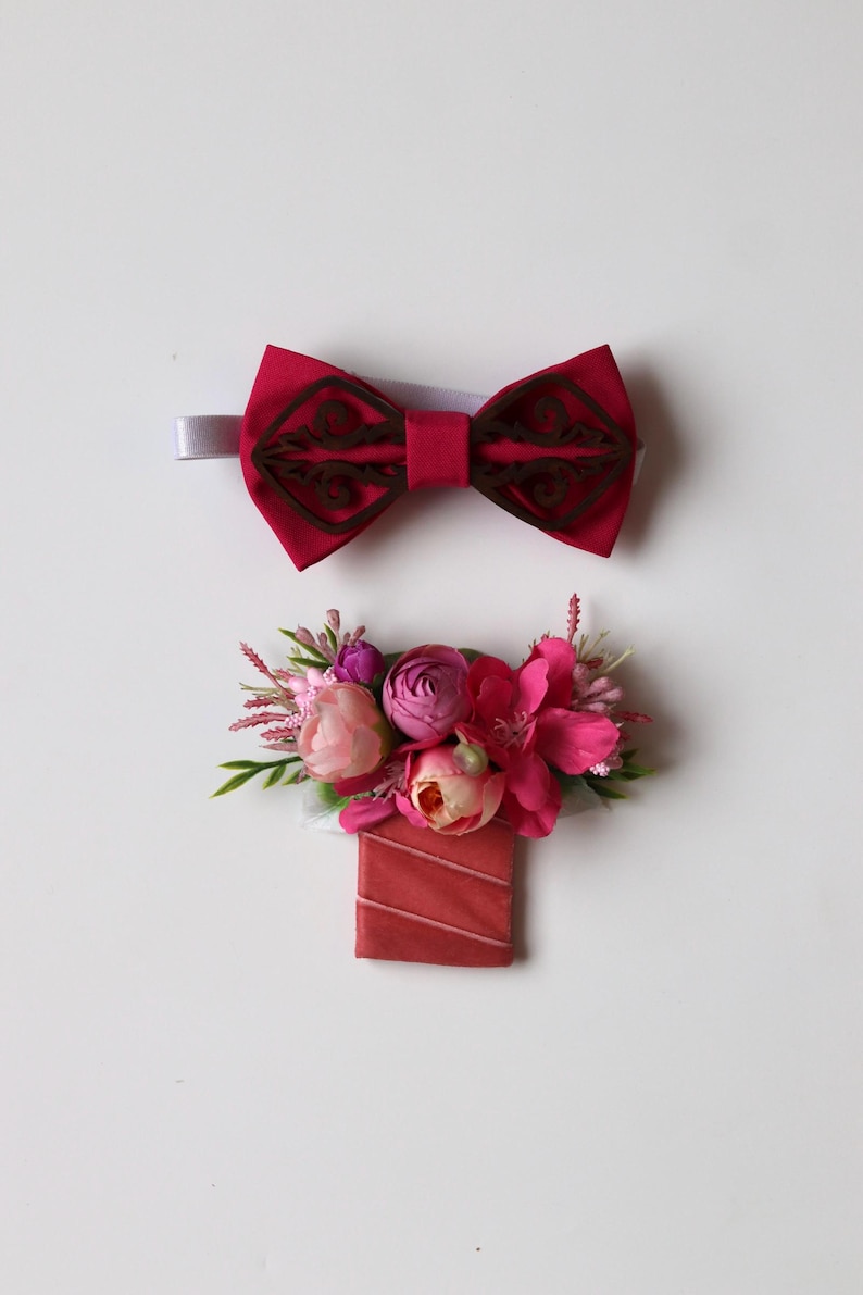 May include: A dark pink fabric bow tie featuring a dark brown laser-cut wooden overlay with intricate scrollwork, paired with a matching boutonniere composed of artificial pink and peach flowers, green foliage, and a rectangular fabric base.