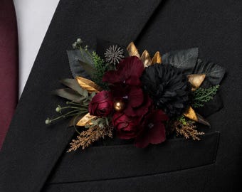 May include: A floral pocket square accessory featuring deep burgundy and black artificial flowers, accented with metallic gold leaves, green foliage, and a small gold bead, tucked into the breast pocket of a black suit jacket with a white shirt.