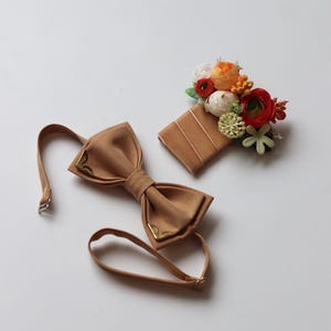 May include: A tan bow tie with gold-toned metal corner accents and an adjustable strap, paired with a matching tan rectangular pocket square adorned with a cluster of artificial flowers in shades of orange, red, white, and green.
