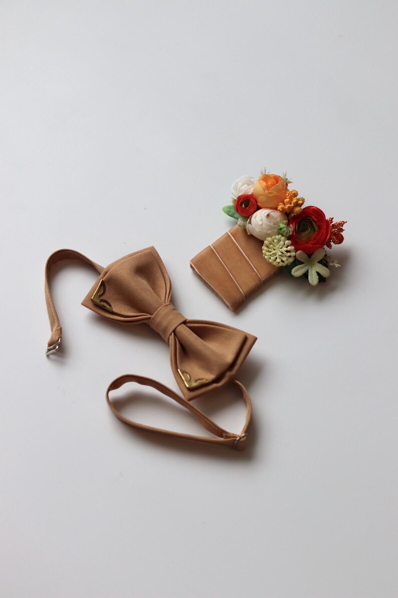 May include: A tan bow tie with gold-toned metal corner accents and an adjustable strap, paired with a matching tan rectangular pocket square adorned with a cluster of artificial flowers in shades of orange, red, white, and green.