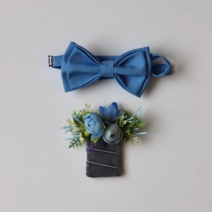 May include: A blue bow tie with an adjustable strap and a matching boutonniere featuring blue fabric flowers, green foliage, and small white berries attached to a rectangular dark gray velvet base with thin silver diagonal ribbon accents.