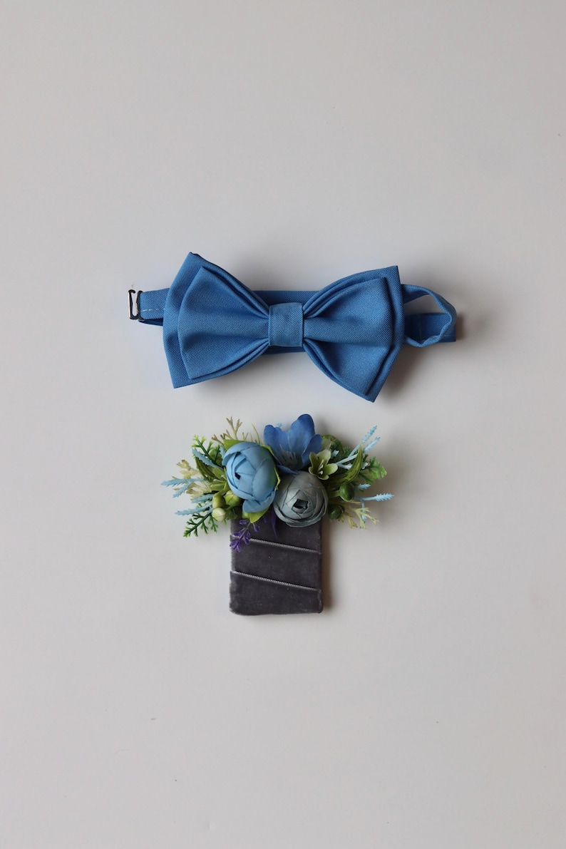 May include: A blue bow tie with an adjustable strap and a matching boutonniere featuring blue fabric flowers, green foliage, and small white berries attached to a rectangular dark gray velvet base with thin silver diagonal ribbon accents.