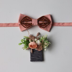 May include: A peach-colored fabric bow tie featuring a dark brown laser-cut wooden overlay with an intricate scrollwork pattern, paired with a boutonniere composed of artificial peach and white flowers, green foliage, and a dark gray velvet ribbon wrap.