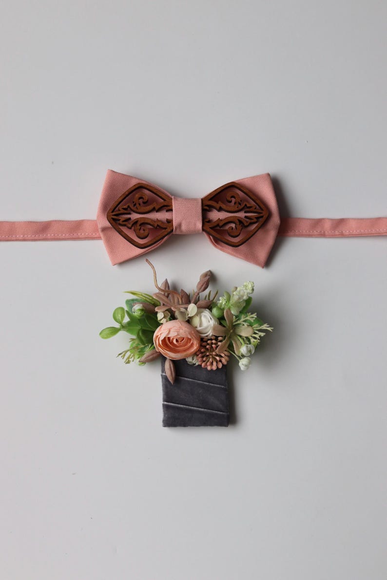 May include: A peach-colored fabric bow tie featuring a dark brown laser-cut wooden overlay with an intricate scrollwork pattern, paired with a boutonniere composed of artificial peach and white flowers, green foliage, and a dark gray velvet ribbon wrap.