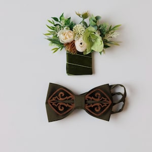 May include: A dark olive green fabric bow tie featuring two symmetrical dark brown laser-cut wooden overlays with intricate scrollwork patterns. Accompanying the bow tie is a matching boutonniere wrapped in dark green velvet ribbon.