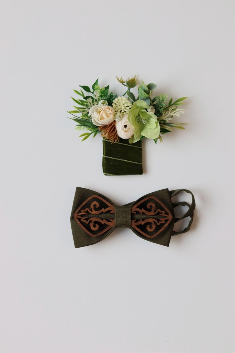 May include: A dark olive green fabric bow tie featuring two symmetrical dark brown laser-cut wooden overlays with intricate scrollwork patterns. Accompanying the bow tie is a matching boutonniere wrapped in dark green velvet ribbon.