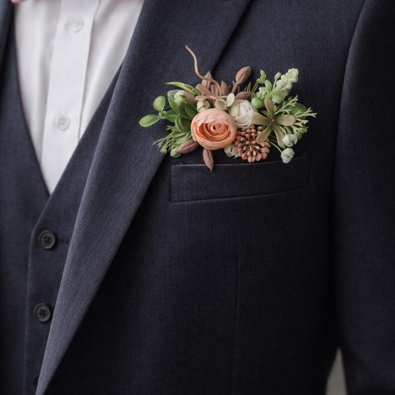 May include: A dark navy blue suit jacket and vest worn over a white button-down shirt. A floral boutonniere featuring a peach-colored rose, small white flowers, green leaves, and brown textured accents is tucked into the jacket breast pocket.